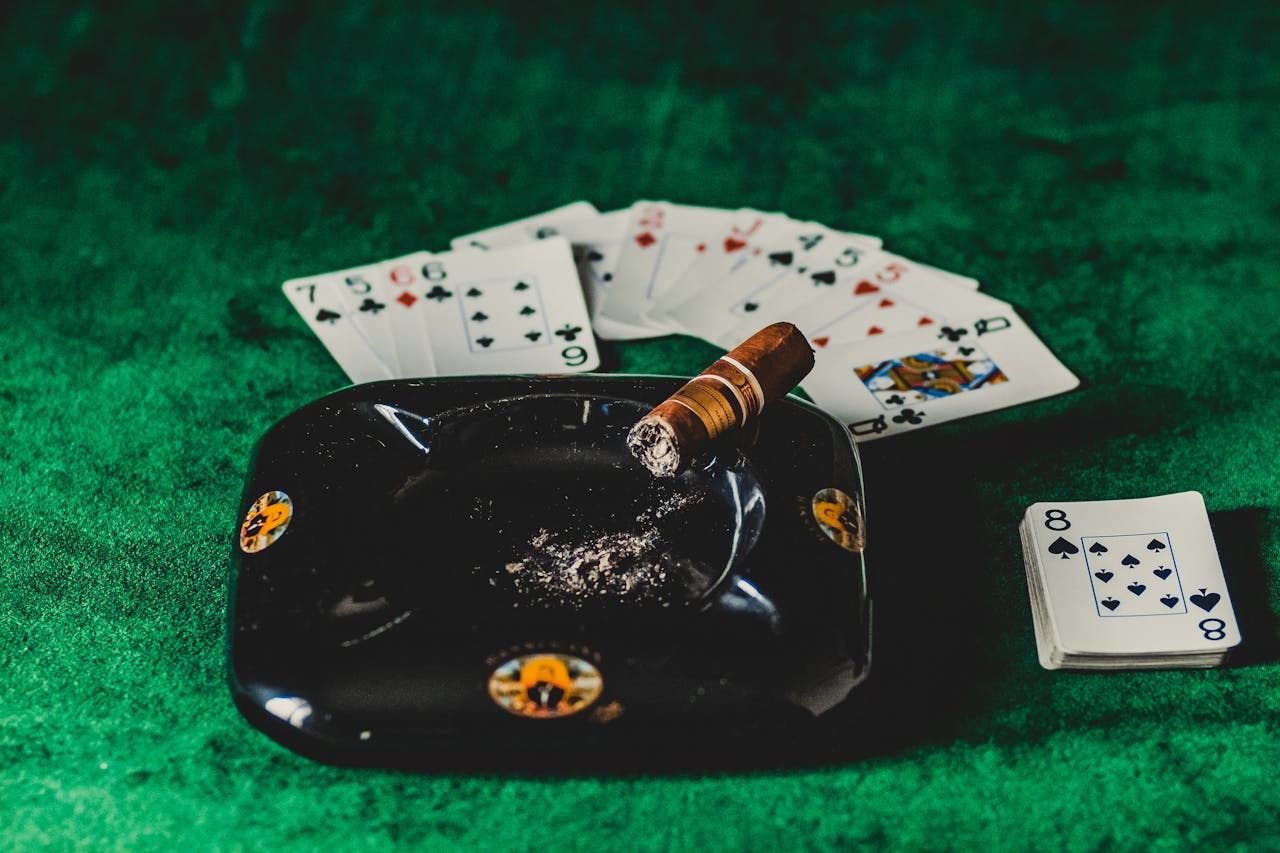 A poker table with playing cards, a lit cigar, and an ashtray on a green felt surface.